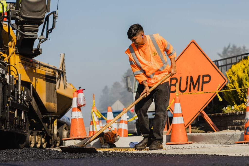 Crew member raking fresh asphalt beside a paver